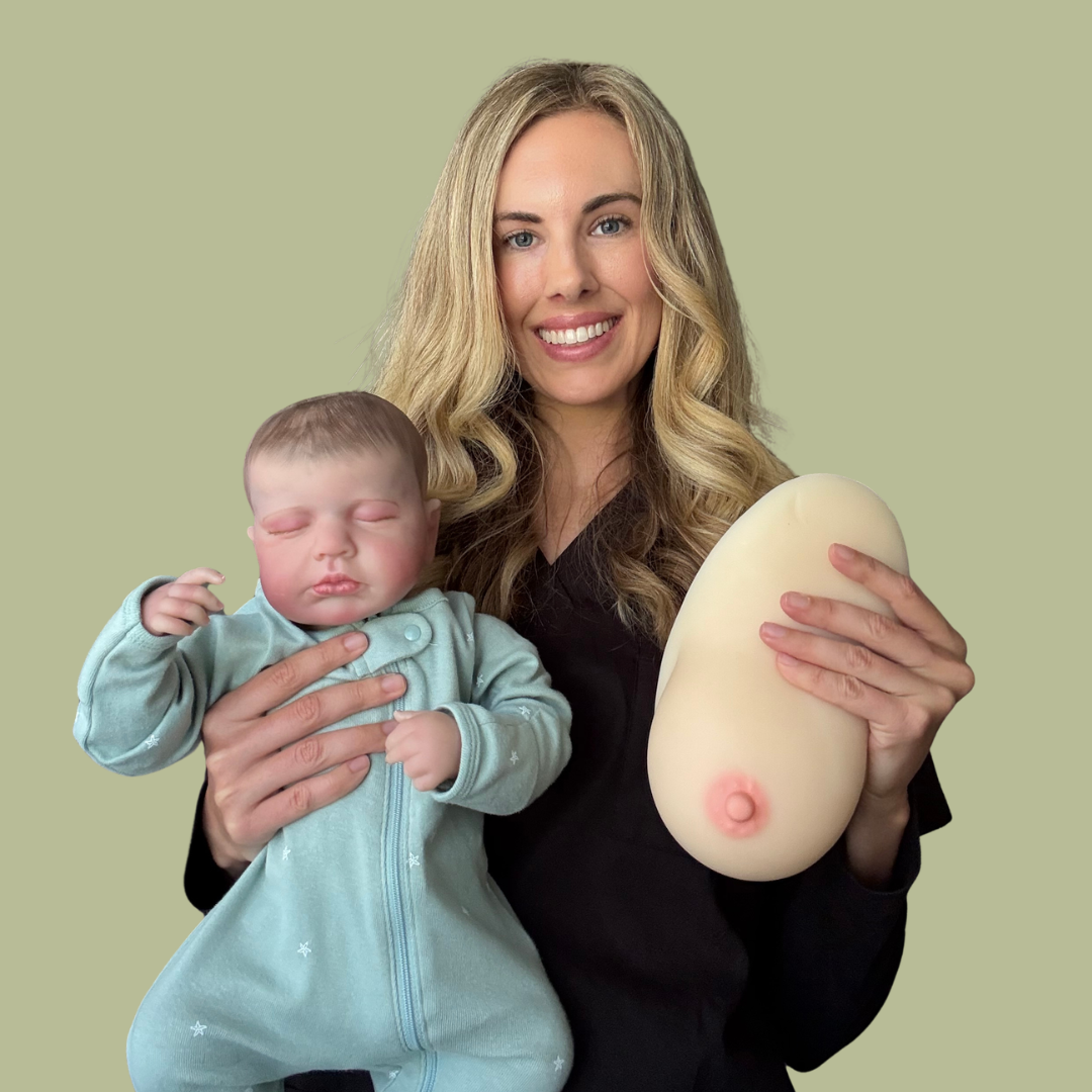 A woman holding a sleeping baby and a large silicone breast mold with a nipple, standing against a light green background.