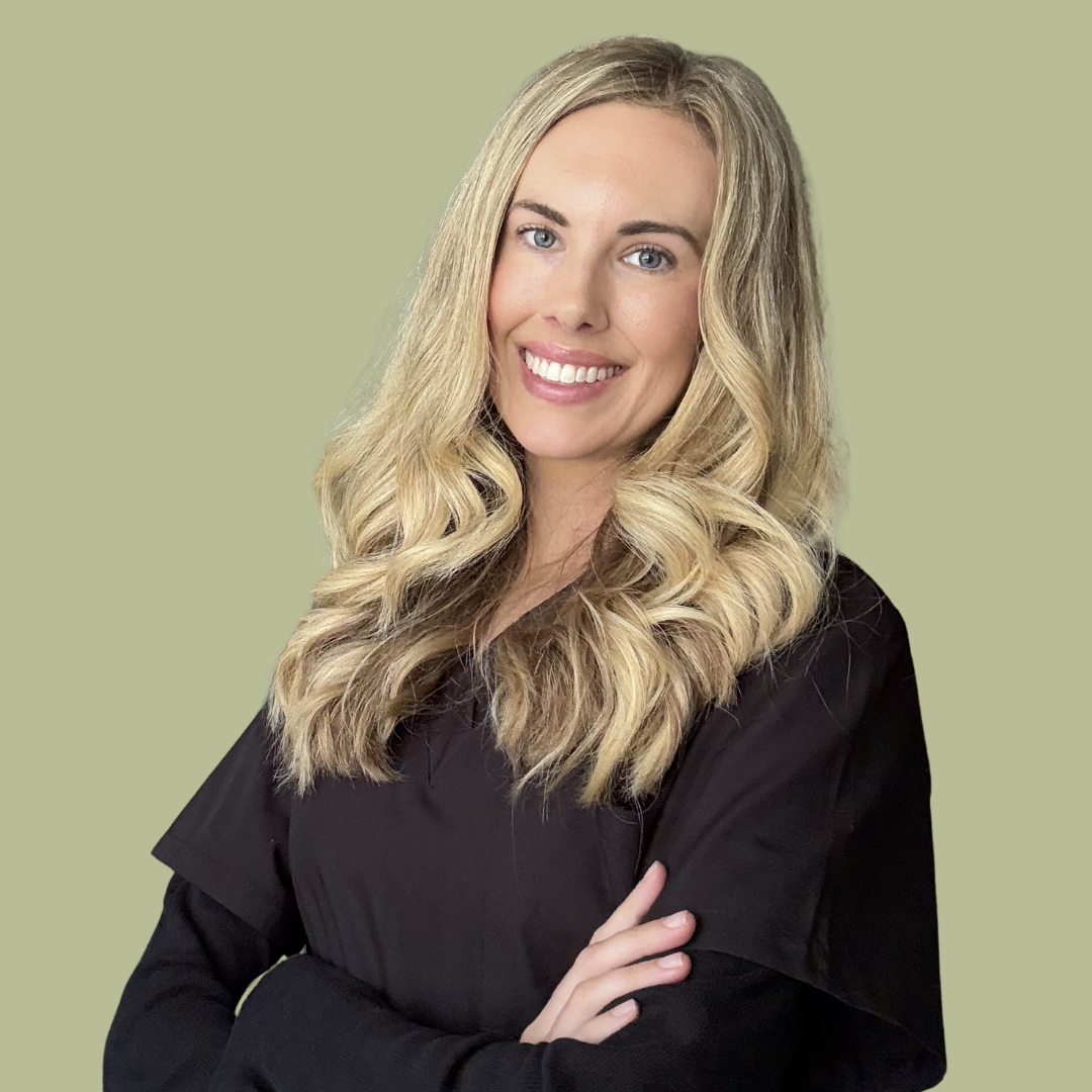 A woman with long blonde wavy hair wearing a black top, smiling with arms crossed, against a plain green background.
