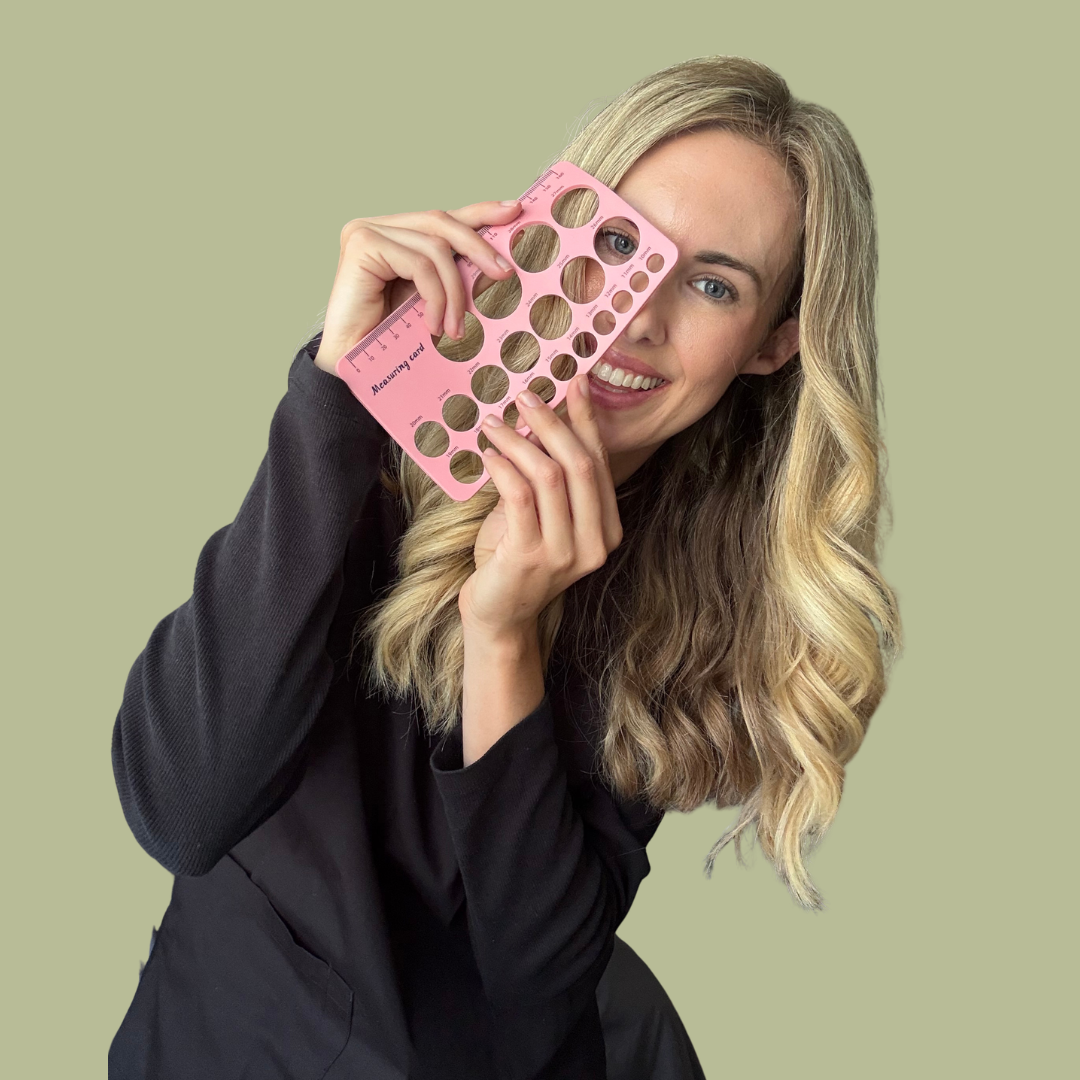 Woman with wavy blonde hair holding a pink measuring template up to her face, smiling, against a solid light green background.