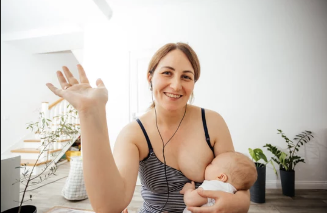 A woman with brown hair wearing a striped tank top is breastfeeding a baby while waving at the camera and smiling.