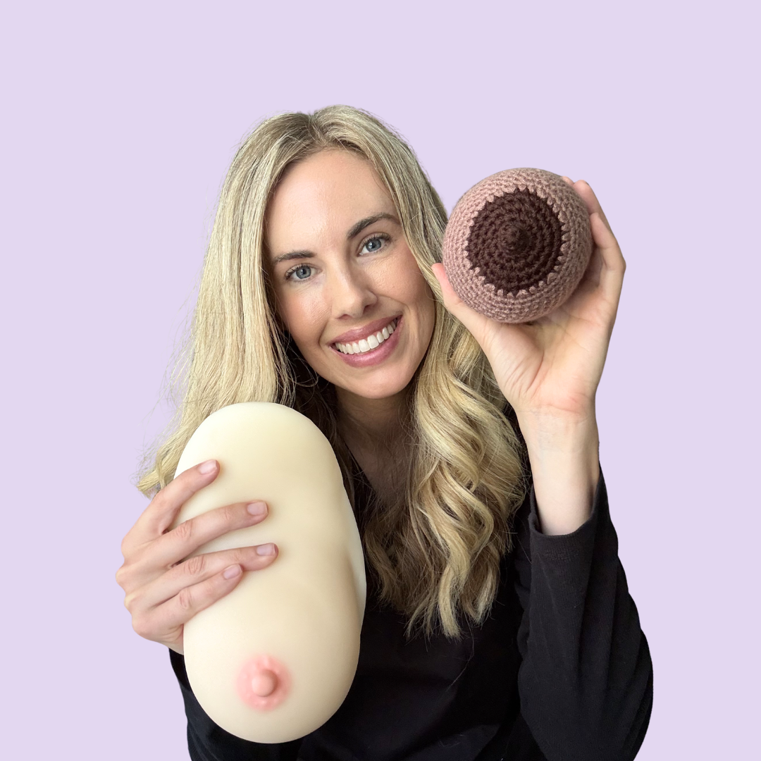 A smiling woman with long blonde hair holding a large, realistic-looking breast model in one hand and a crocheted egg in the other, against a light purple background.