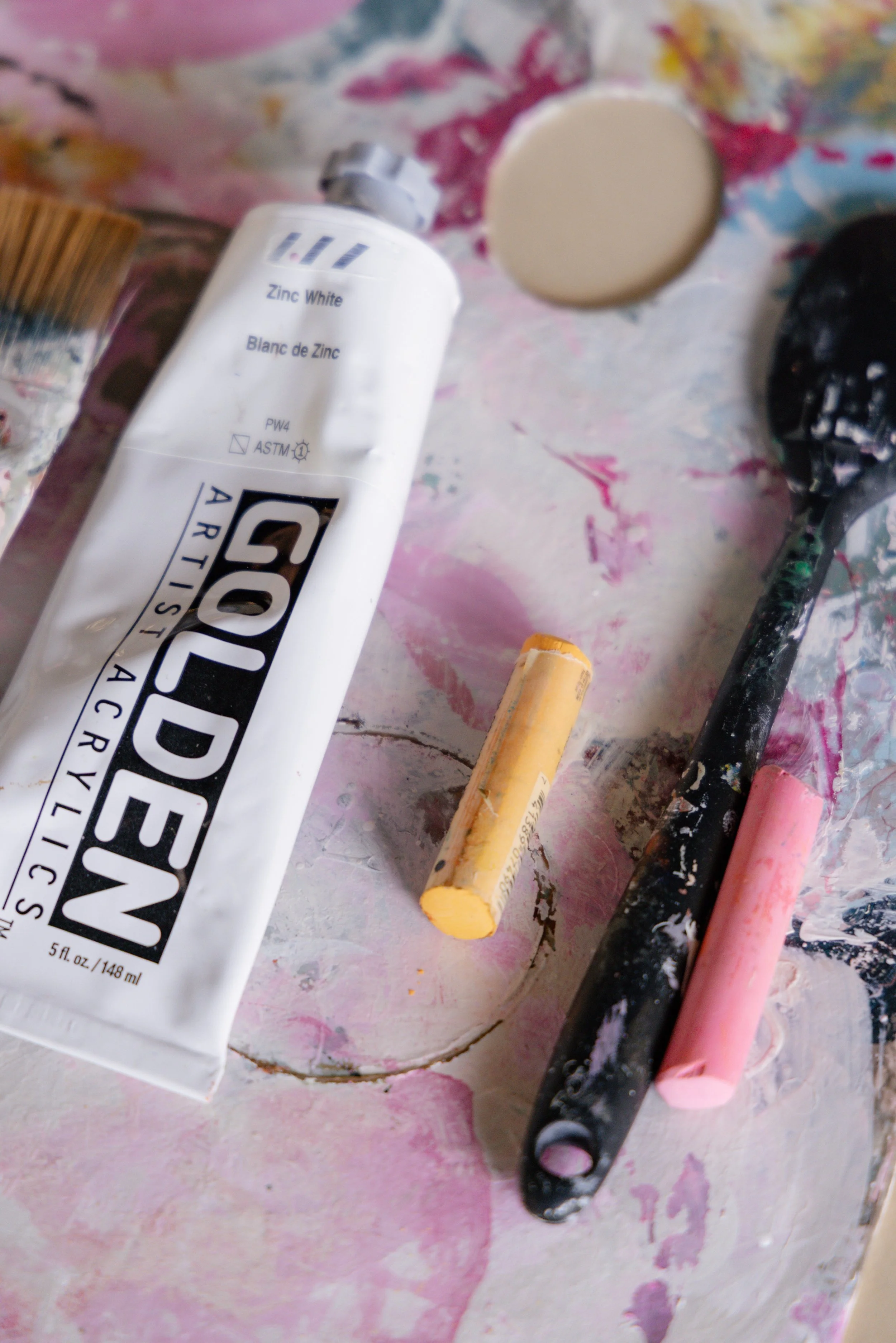 A tube of white acrylic paint, a paint palette with pink and beige paint, and some paint-dipped brushes on a surface with pink and purple paint splatters.