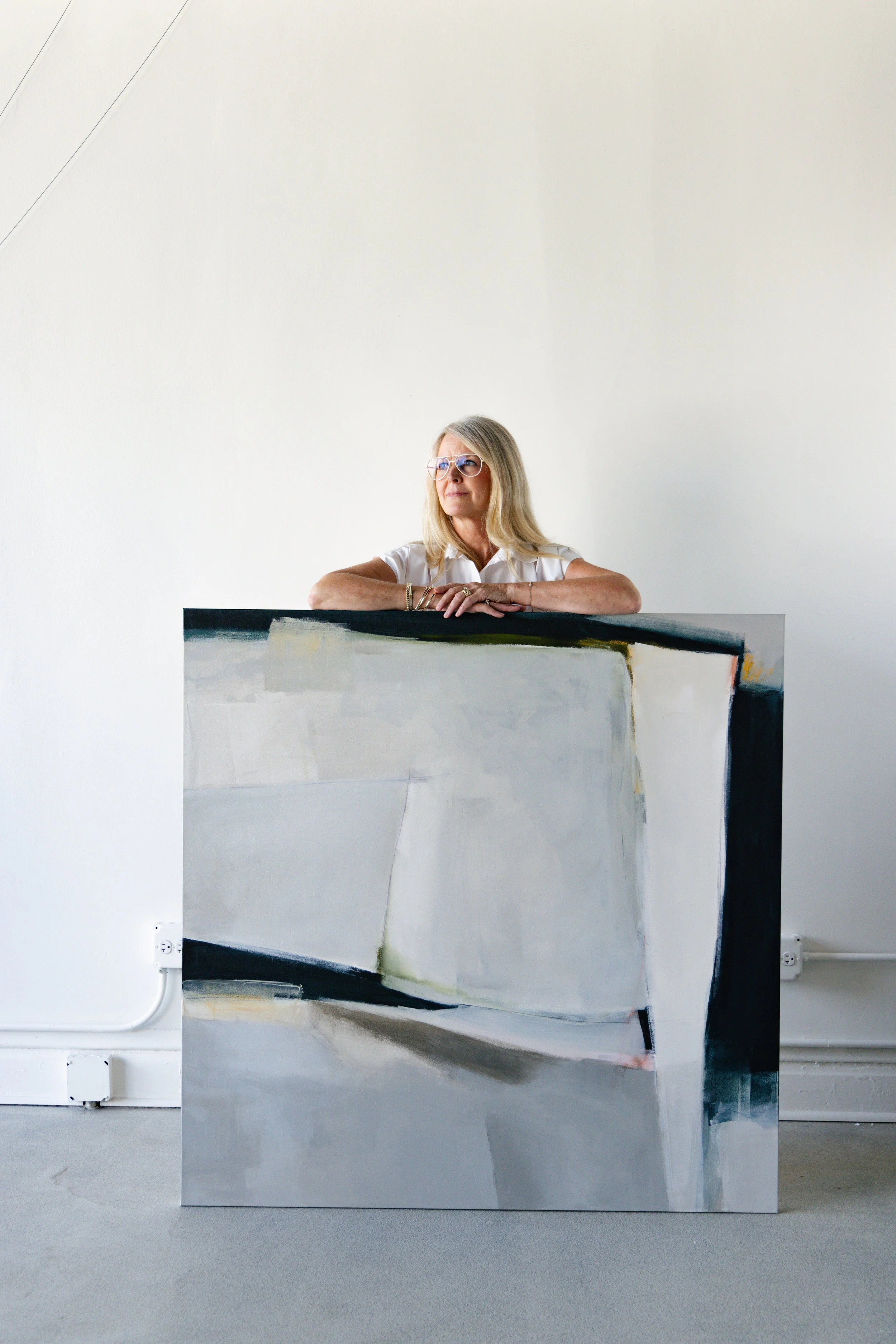 A woman with blonde hair and glasses leaning on a large abstract painting in an art gallery or studio.