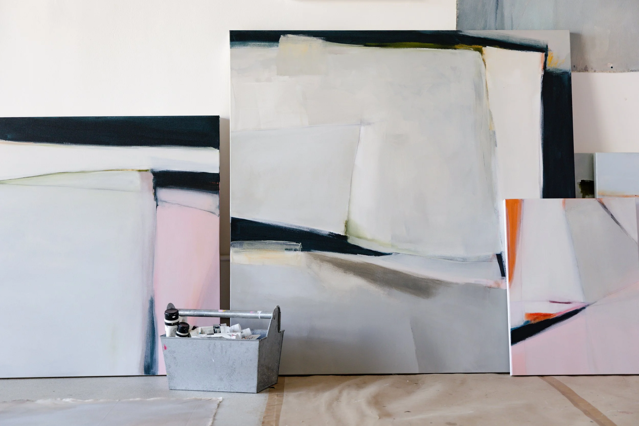 Abstract paintings leaning against a wall with painting supplies in a container in front.