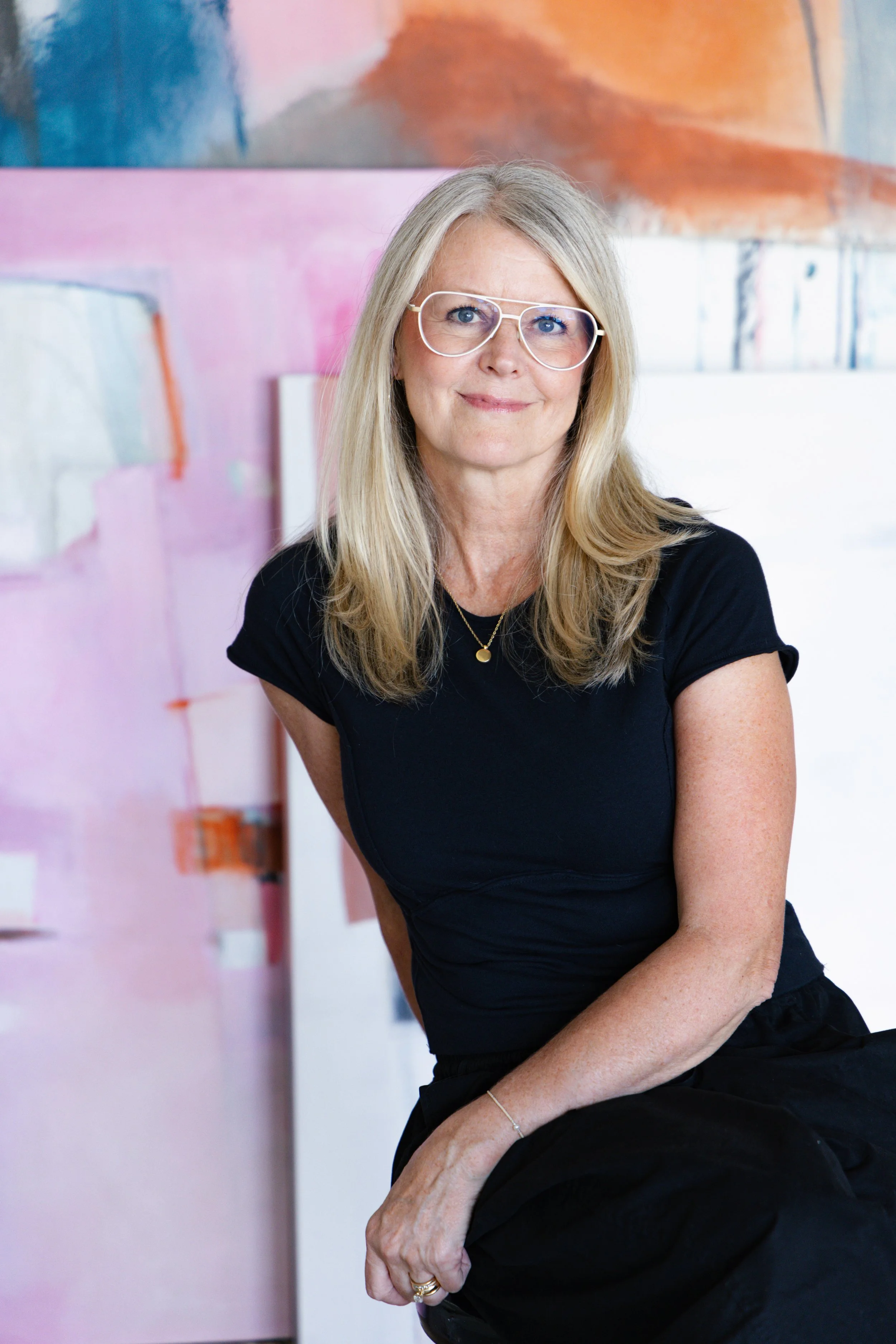 Portrait of a woman with long blonde hair, wearing glasses and a black shirt, sitting in front of an abstract colorful painting.