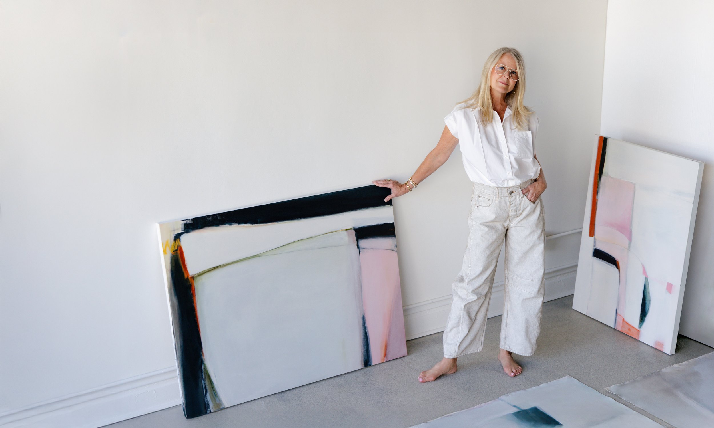 A woman with long blond hair, glasses, white short-sleeved shirt, and beige pants standing barefoot in an art studio, leaning on a large abstract painting resting against a white wall, with additional paintings on the floor.
