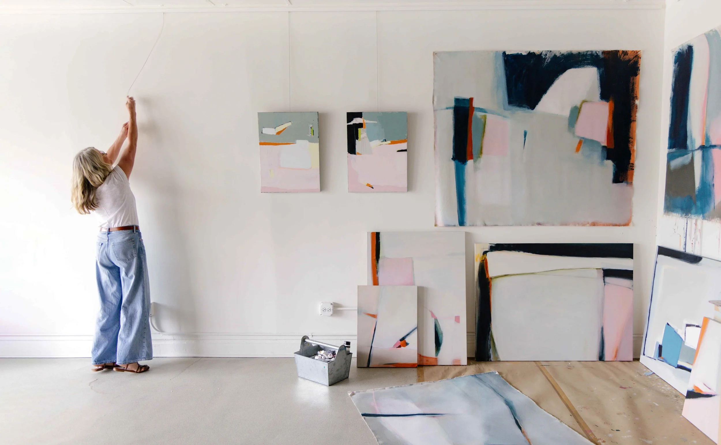 A woman with blonde hair, glasses, white shirt, and blue jeans is hanging a wire on a white wall in an art studio. Several abstract paintings with pastel, black, and orange colors are displayed on the wall and on the floor, with some leaning against the wall, and a small metal container with art supplies is on the floor.