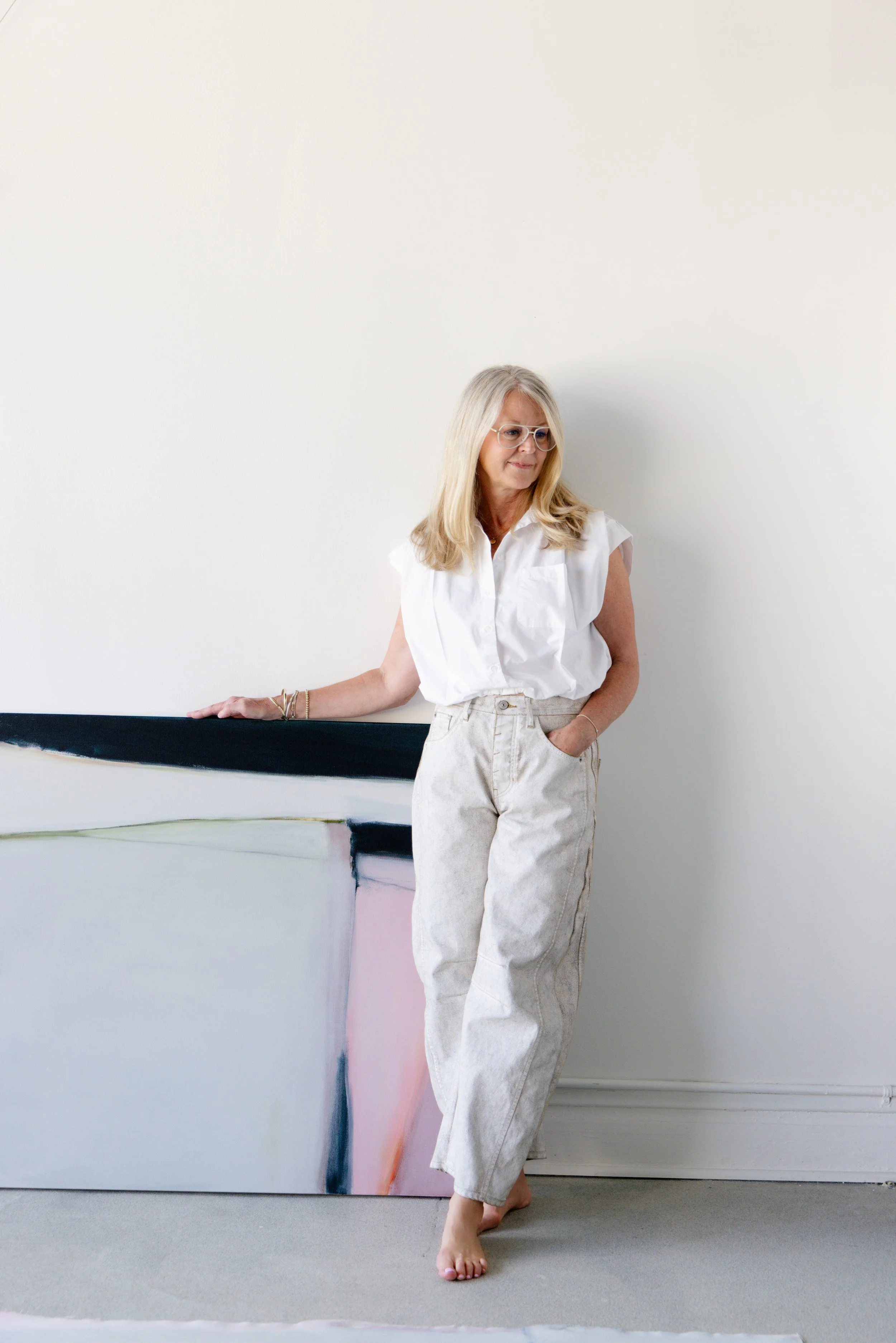 A woman with blonde hair wearing glasses, a white sleeveless blouse, and beige pants standing barefoot in front of an abstract painting with pastel colors, leaning on the edge of the painting with her left hand.
