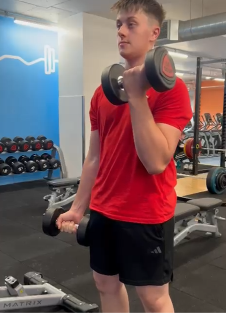 Young man in a red t-shirt and black shorts lifting dumbbells in a gym.