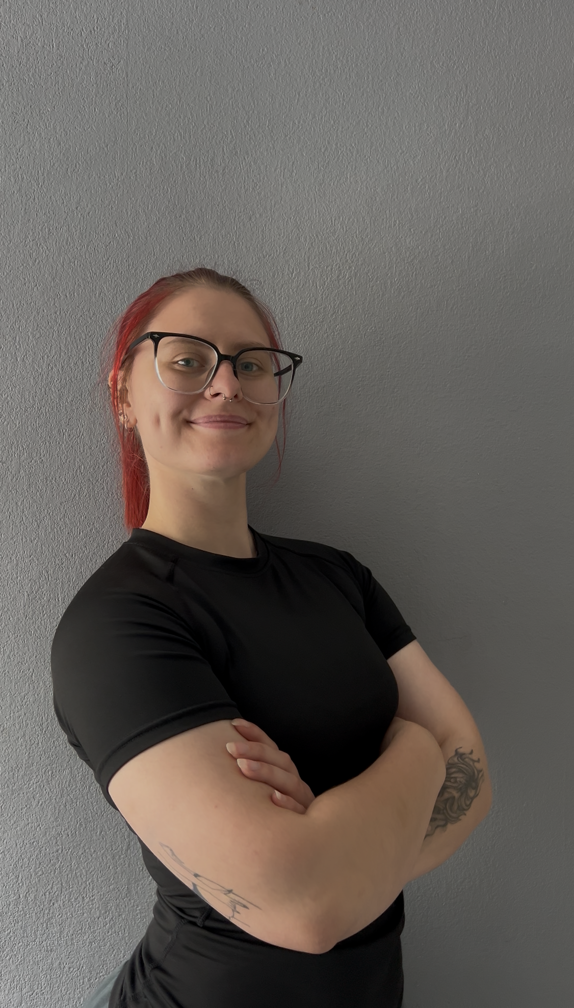 A woman with red hair, glasses, and tattoos on her arms, standing against a gray wall, smiling with her arms crossed.