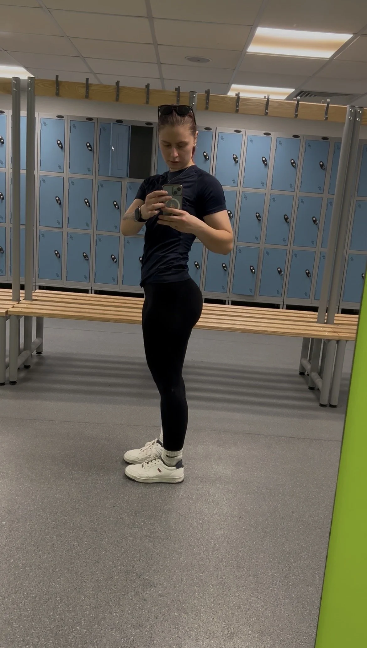A young woman taking a mirror selfie in a locker room, wearing a black t-shirt, black leggings, white sneakers, and sunglasses on her head.