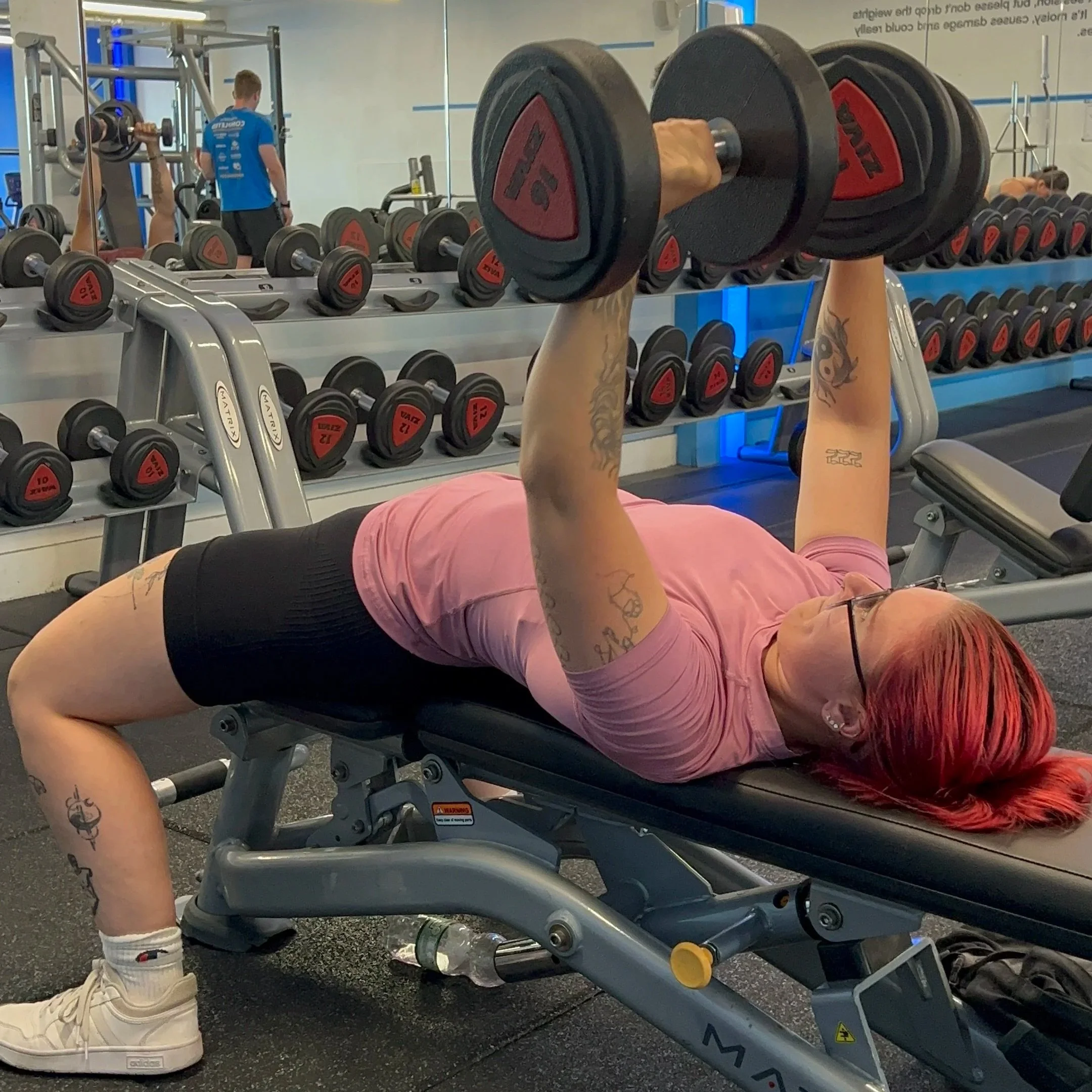 A person lying on a bench in a gym, lifting a dumbbell with arms raised, with a row of various dumbbells behind them.