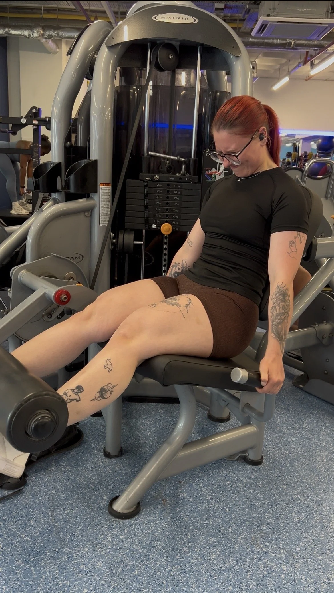 A woman with tattoos is sitting on a leg press machine at the gym, smiling and looking down. She is wearing glasses, a black t-shirt, and brown shorts. There are weight stacks and other gym equipment around her.