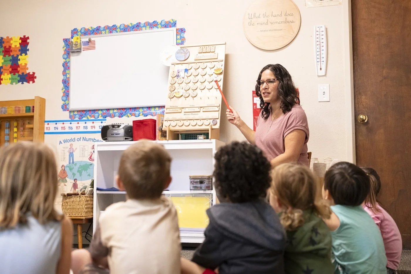 A teacher standing in front of a classroom with young students sitting on the floor. The teacher is pointing at a board with a pointer. Educational posters and a calendar are visible on the wall.