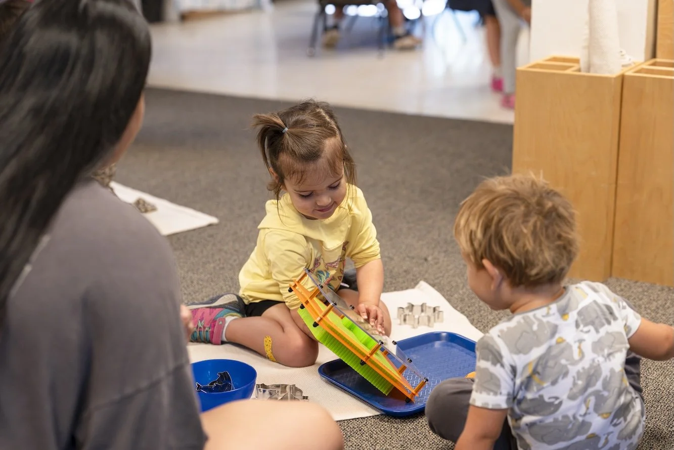 Two young children playing with a colorful marble run toy on the floor while an adult watches. The girl is focused on placing a marble in the toy, and the boy watches attentively. The setting appears to be indoors, likely a classroom or playroom.