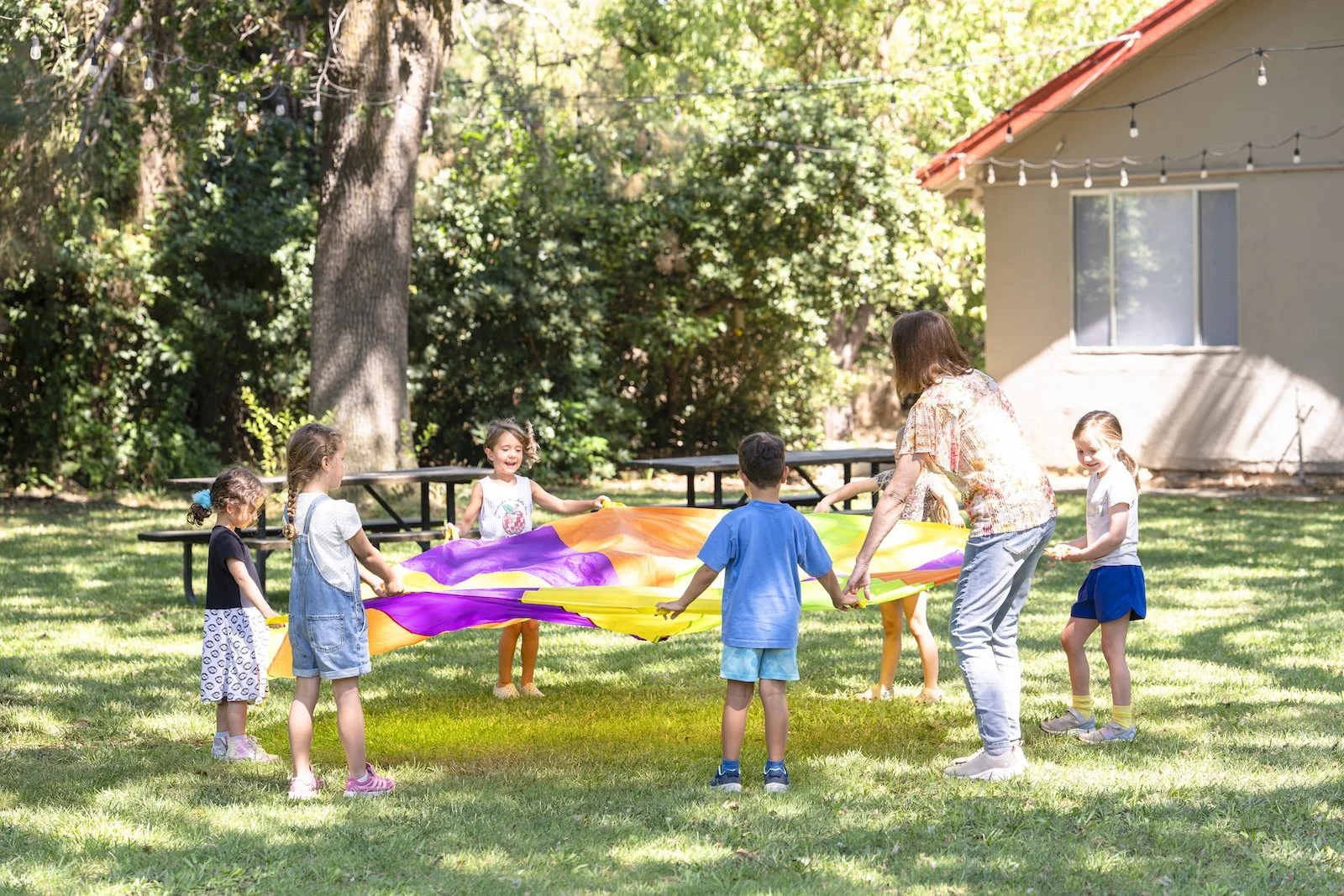 Children and an adult playing with a colorful parachute outdoors in a grassy yard surrounded by trees, near a house with string lights.