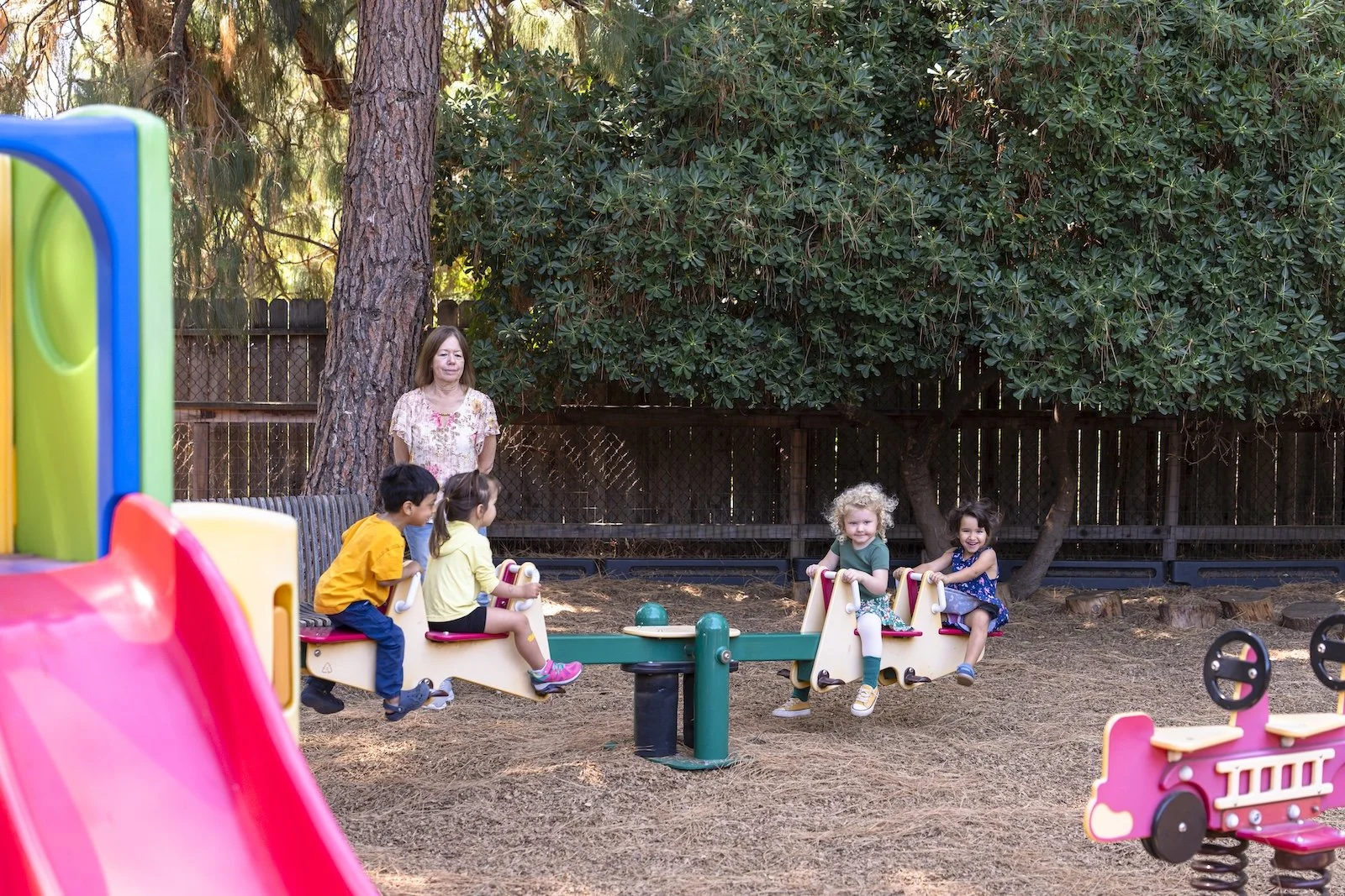 Four children playing on a see-saw in a playground with a woman supervising. There are trees and a wooden fence in the background.