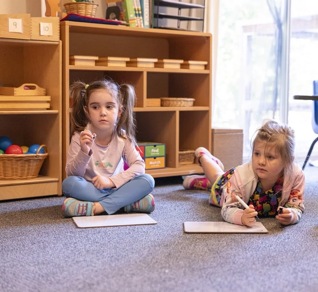Two young girls sitting and lying on the carpeted floor in a classroom, each with a dry erase board and marker, with shelves filled with educational toys and books in the background, near a large window.