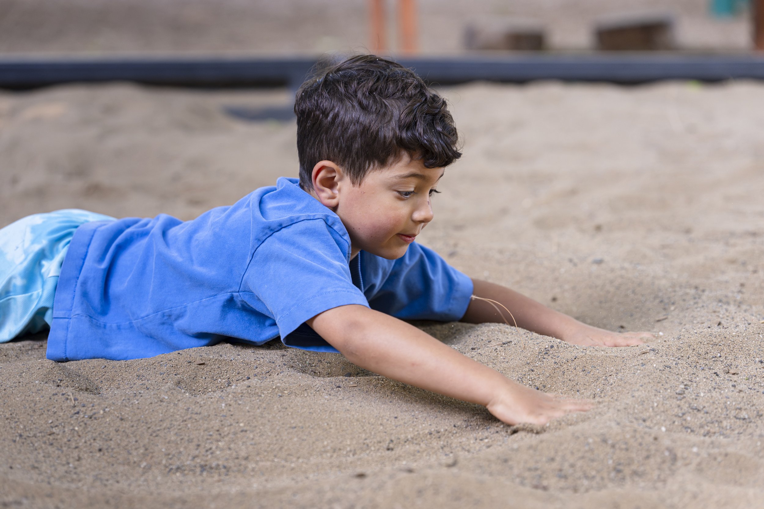 A young boy in a blue shirt lying face down on the sandy ground at the beach, drawing or playing with the sand.