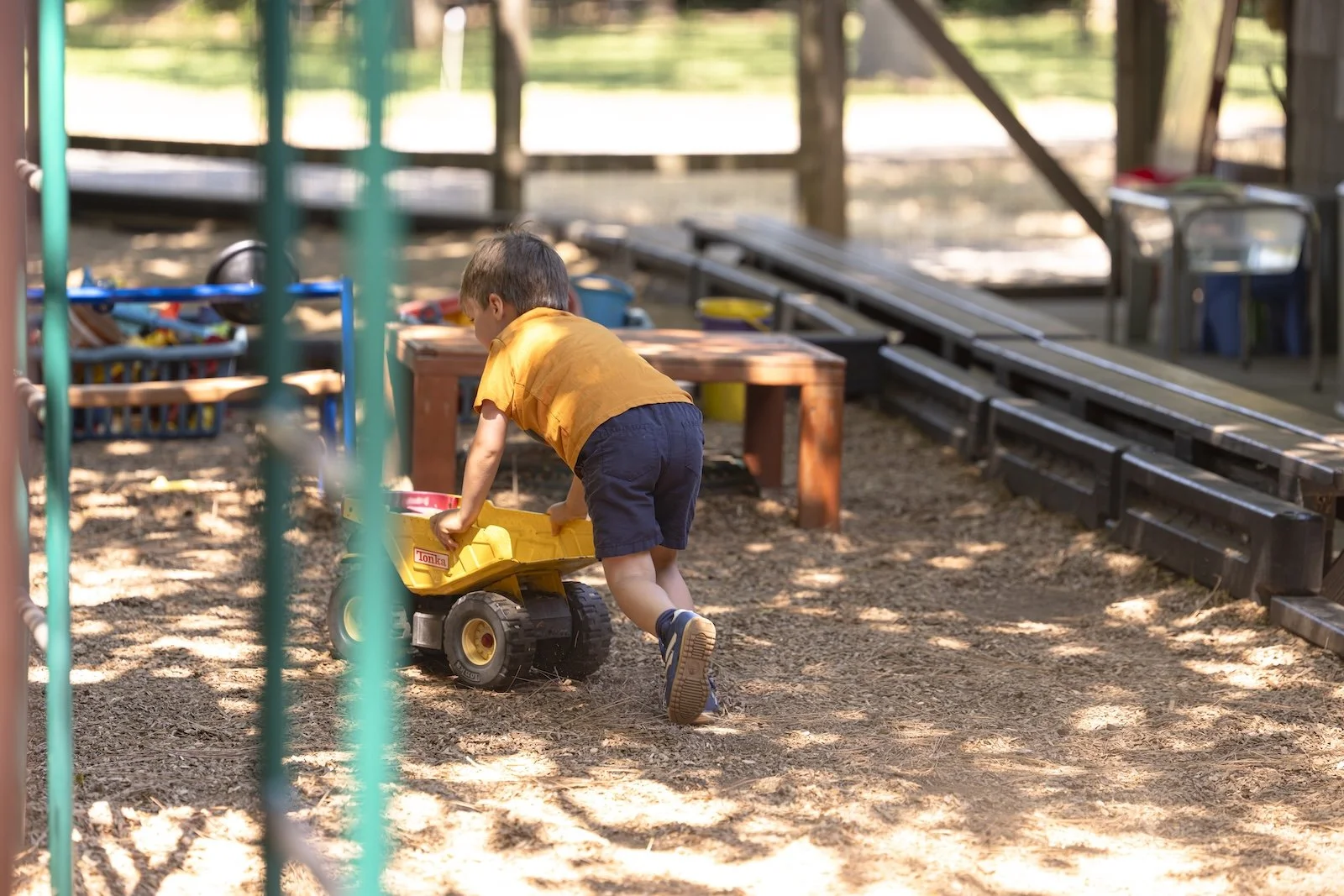 A young boy wearing an orange shirt and navy shorts is playing with a yellow toy dump truck on a dirt playground. There are wooden tables, benches, and various toys around him, with sunlight filtering through the trees.