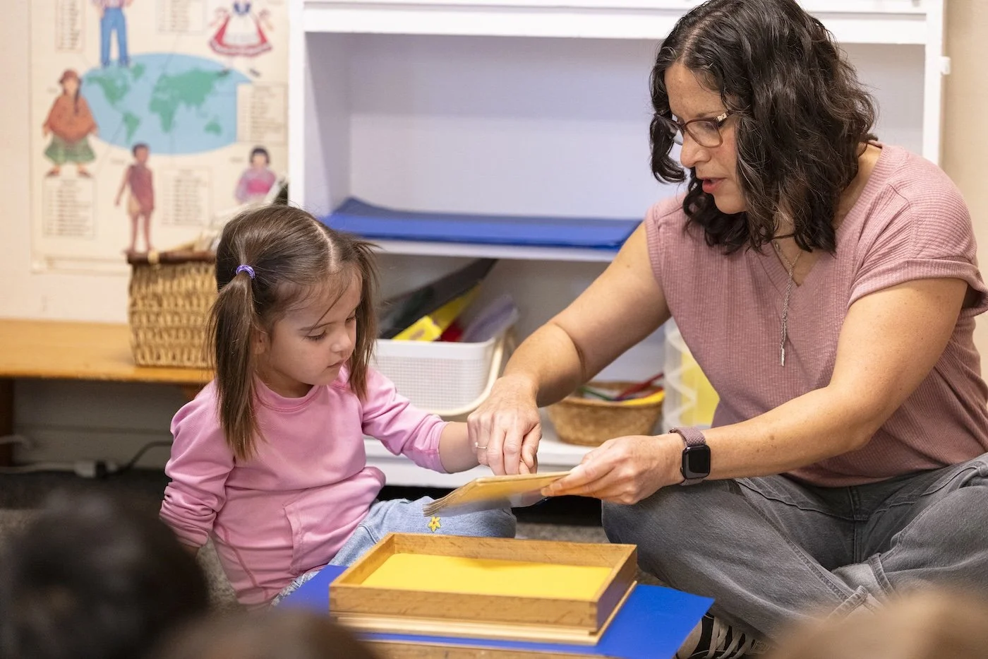 A woman and a young girl sit on the floor in a classroom, engaging with a book or folder together. The woman is wearing glasses and a pink shirt, and the girl has brown hair tied in a ponytail with a purple hair tie, dressed in a pink long-sleeve shi