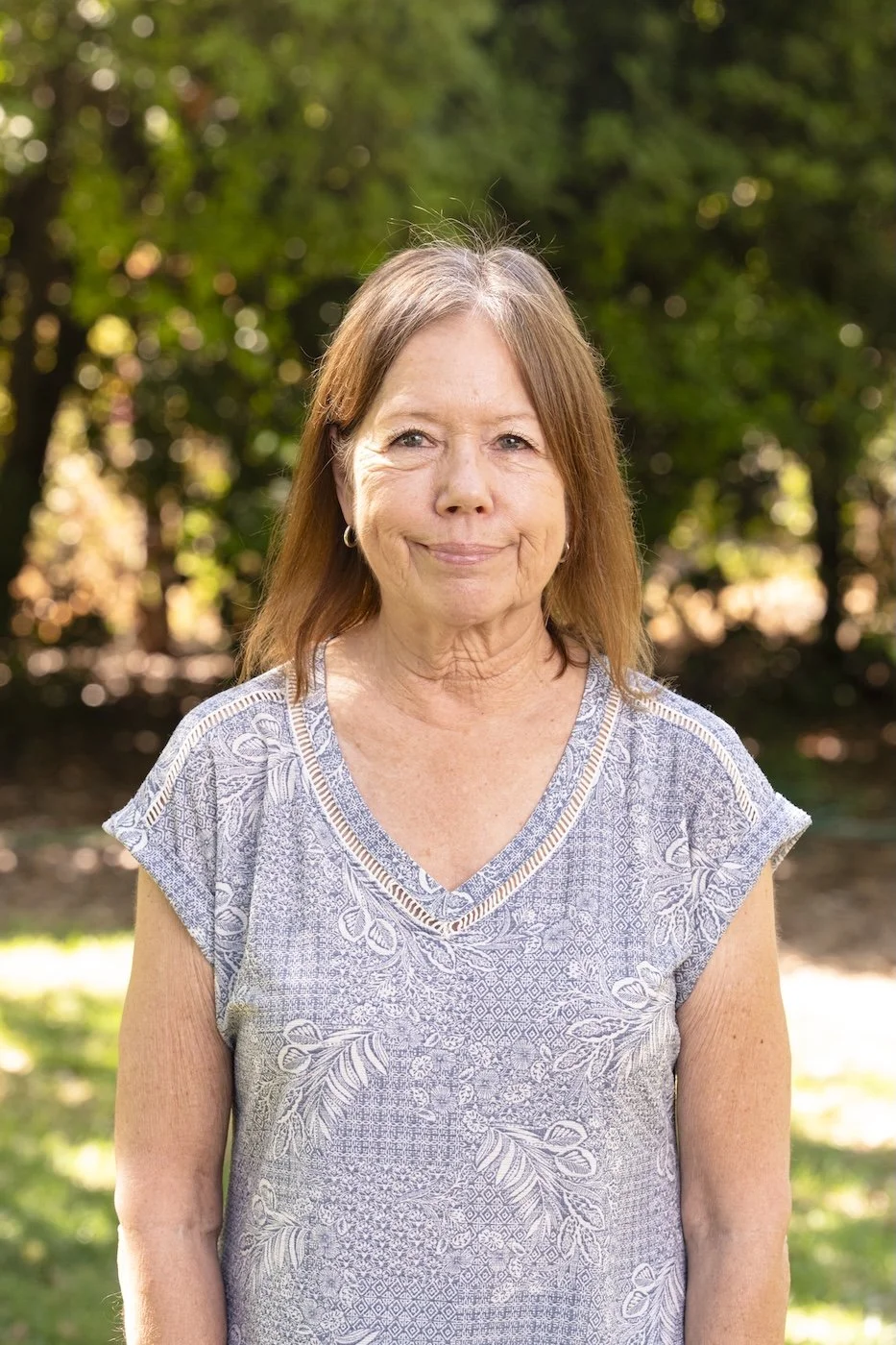 A woman with shoulder-length brown hair and light skin, wearing a gray patterned top, outdoors on a sunny day with green trees and foliage in the background.
