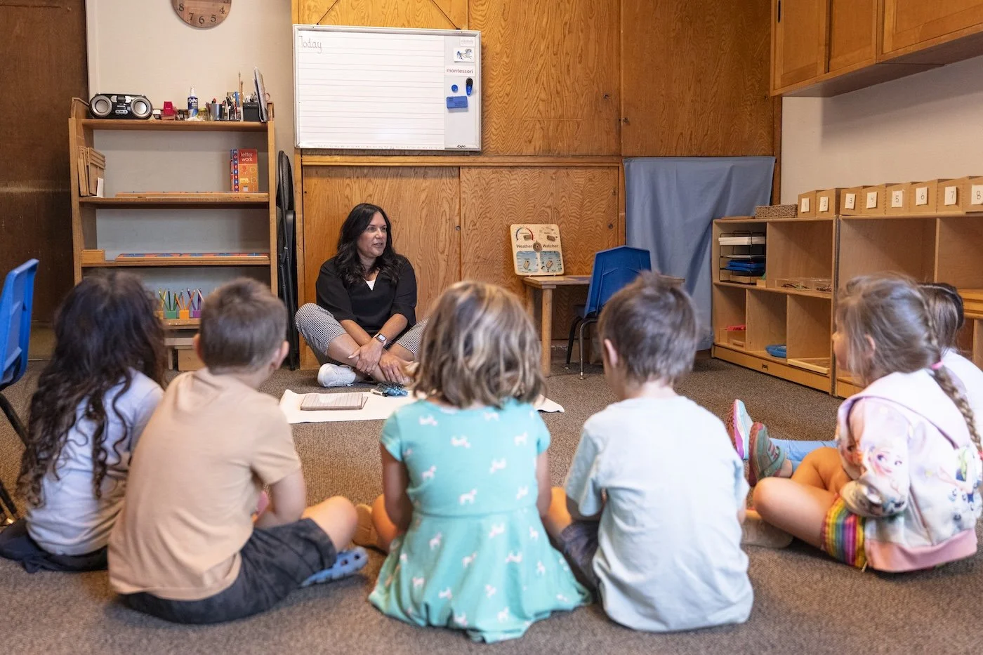 A teacher sitting on the floor with a group of young children sitting on the carpet in a classroom. The background has wooden shelves, a whiteboard, and educational materials.