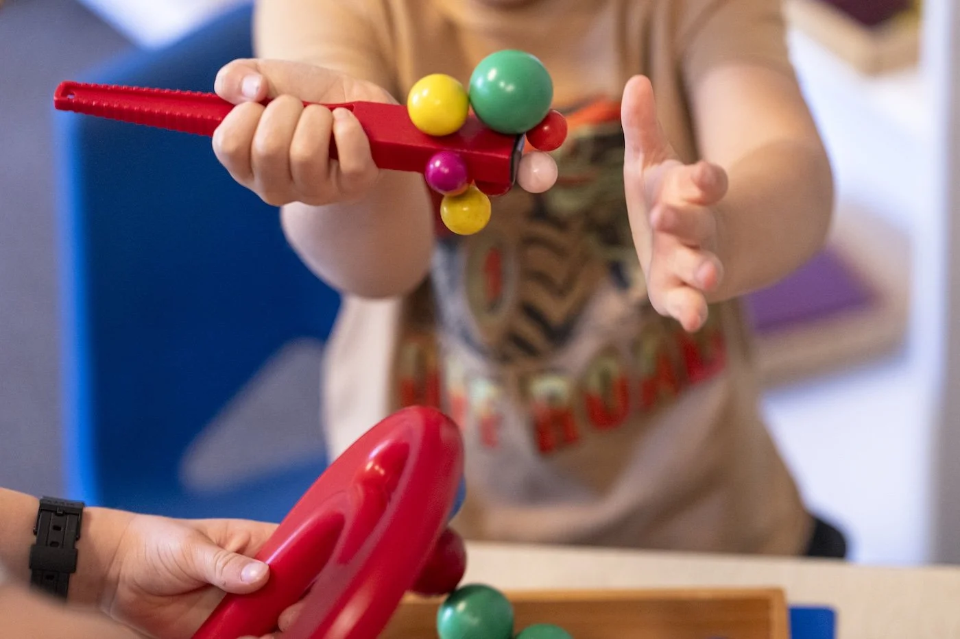 Child playing with a red toy gun with colorful balls attached, held by a caregiver, in a room with a blue chair and a wooden table.