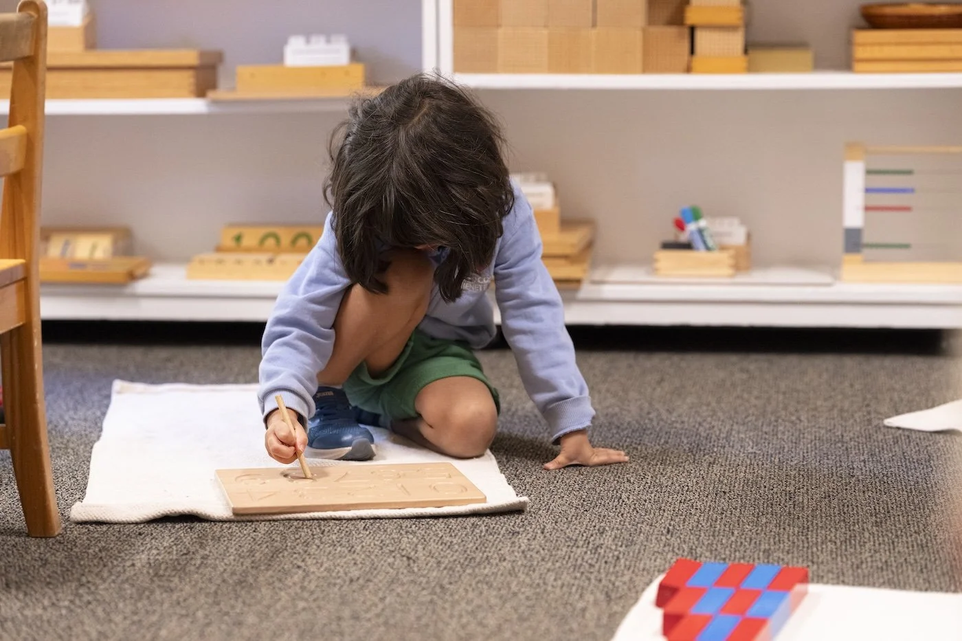 Child kneeling on a white cloth, drawing on a wooden board with a pencil in a classroom setting.