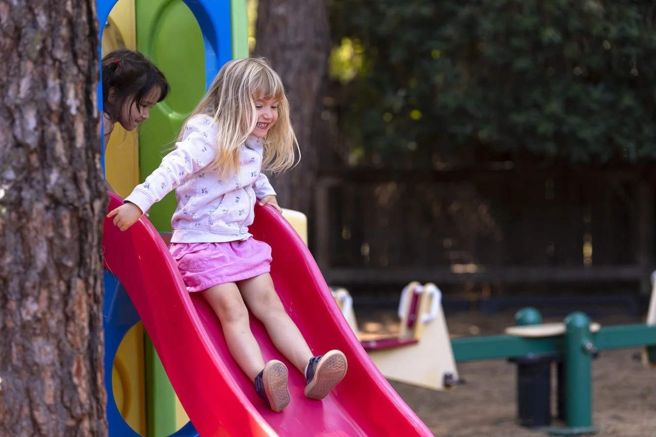 Two young girls playing on a red slide at a playground surrounded by trees.