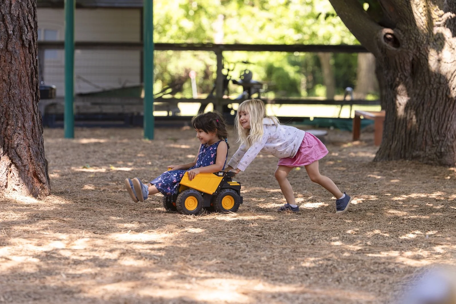 Two young girls playing outdoors with a toy dump truck among trees and shaded ground.