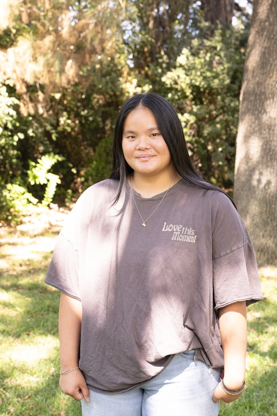 A young woman with long black hair standing outdoors in front of trees and foliage, wearing a loose tie-dye gray T-shirt with text and light gray pants, smiling with her hands in her pockets.