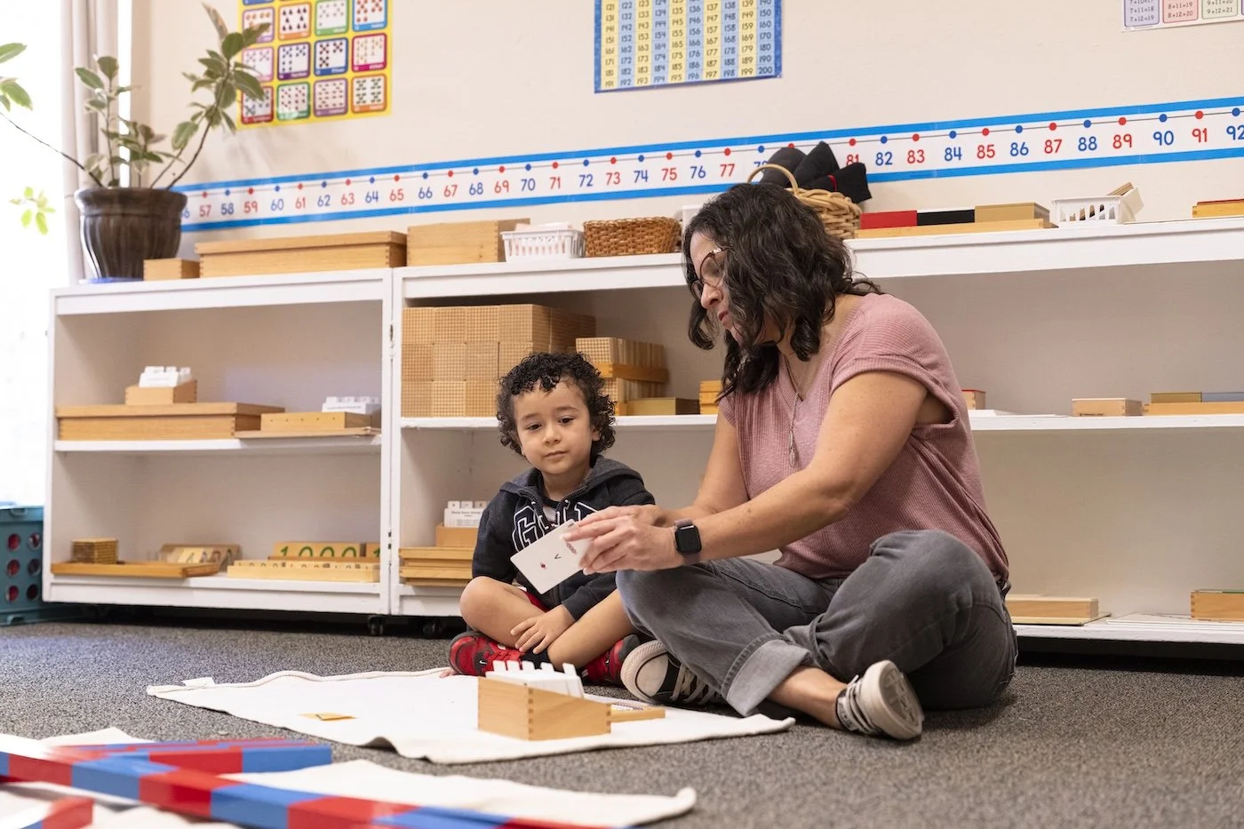 A woman and a young boy sitting on the floor in a classroom, engaging in an educational activity with wooden blocks and cards, with shelves, educational posters, and a plant behind them.