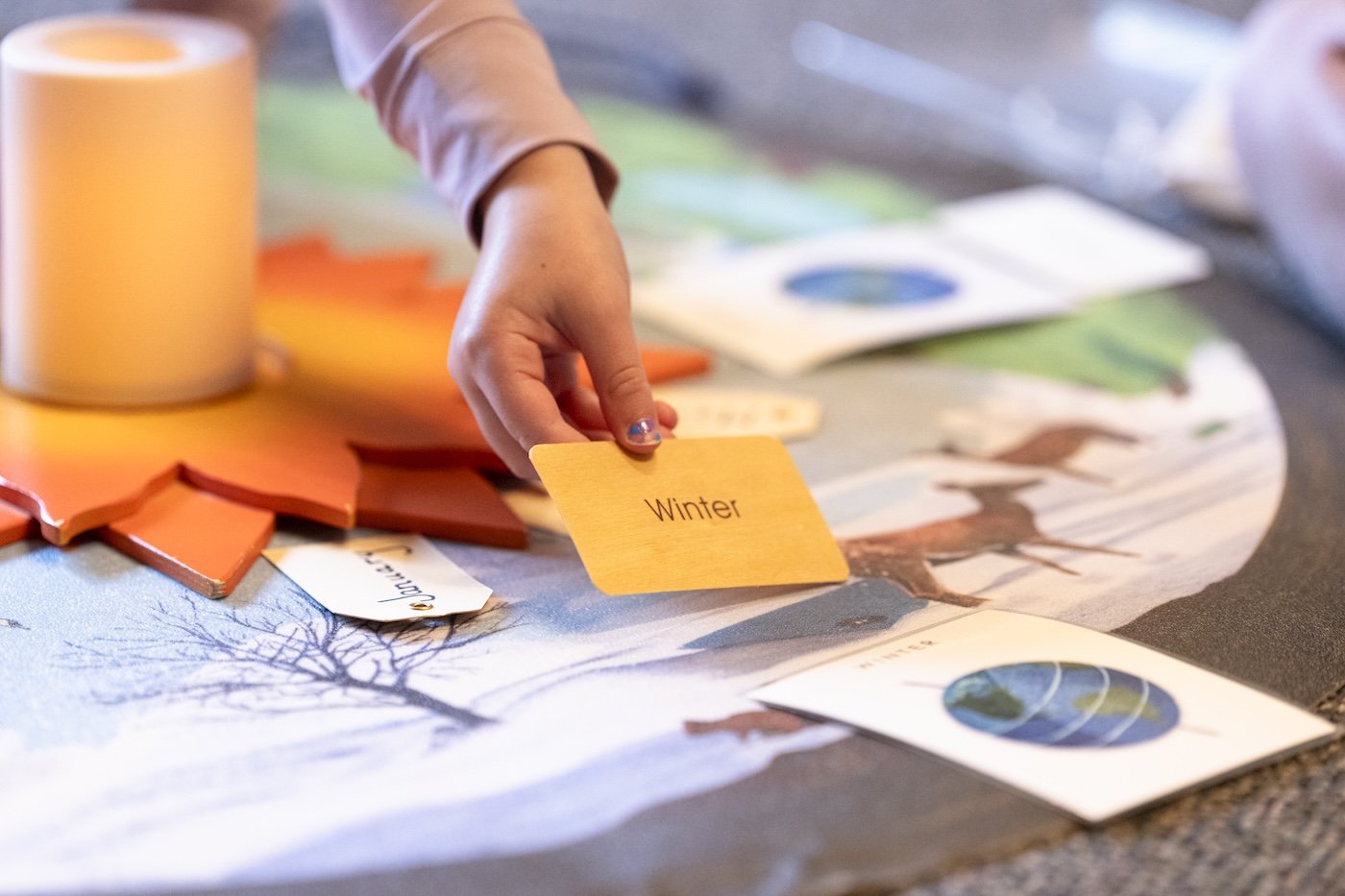 A child's hand holds a yellow card labeled 'Winter' on a table with educational materials, including other cards, a rolled-up paper, and a colorful tree-shaped object, likely for a learning activity about seasons.