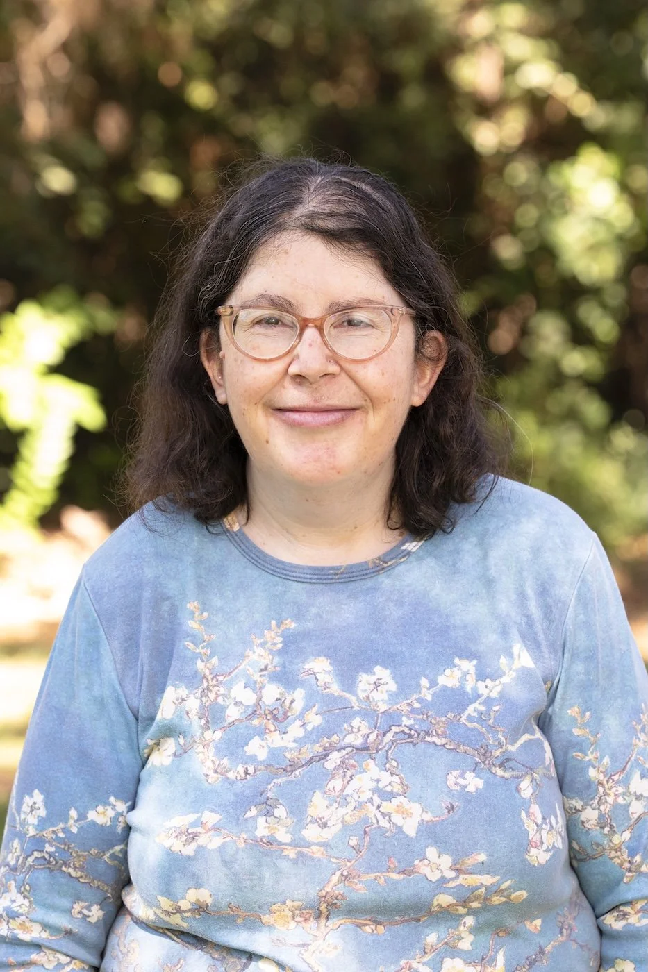 A woman with glasses and dark hair smiling outdoors, wearing a blue sweatshirt with a floral pattern, with a blurred background of trees.