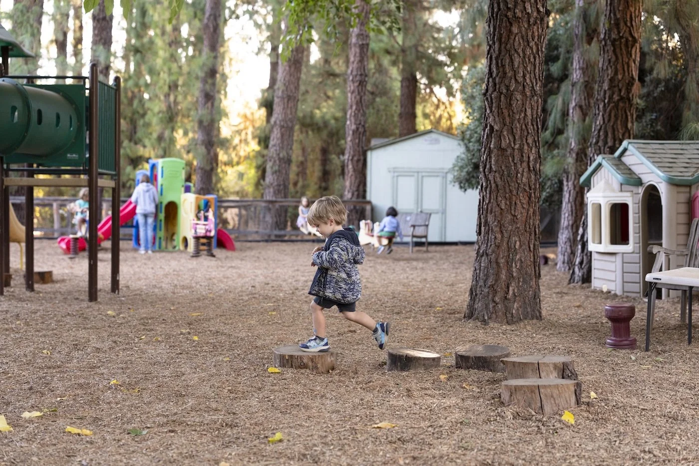 Child jumping over tree stumps in a park with playground equipment and trees in the background.