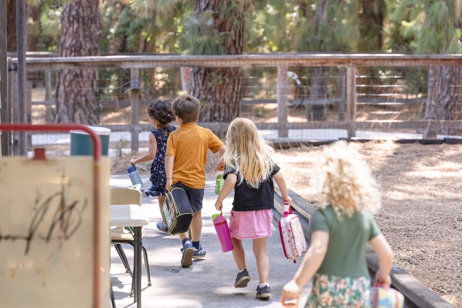 Children walking with backpacks and lunch boxes in an outdoor setting, possibly at a school or park, with trees and fencing in the background.