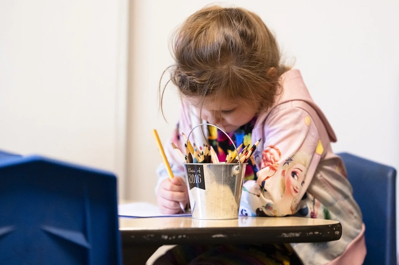 A young girl sitting at a table, leaning forward, drawing or coloring with colored pencils from a metal container labeled "Class of 2016."