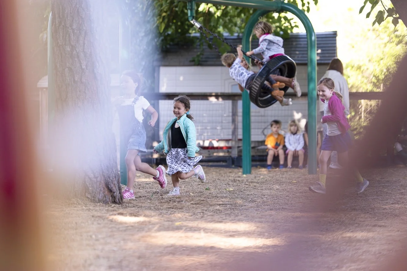 Children playing on a playground swing set, some running on the ground, and others sitting on a bench in the background, with trees and a shed nearby.