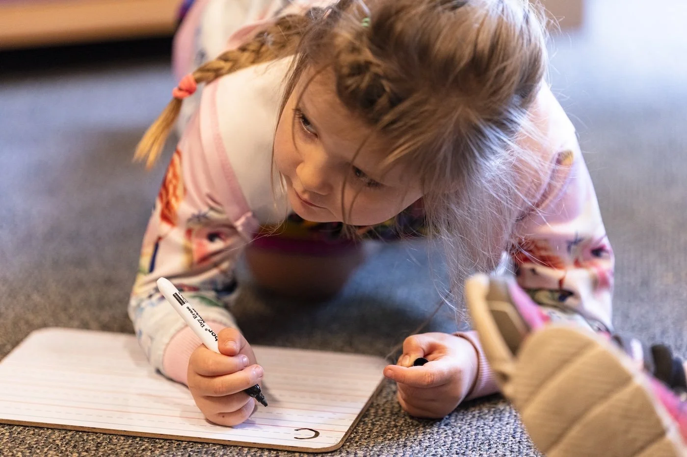 A young girl with braided hair is lying on the carpeted floor, writing on a white-lined notepad with a black marker. She appears focused on her writing.
