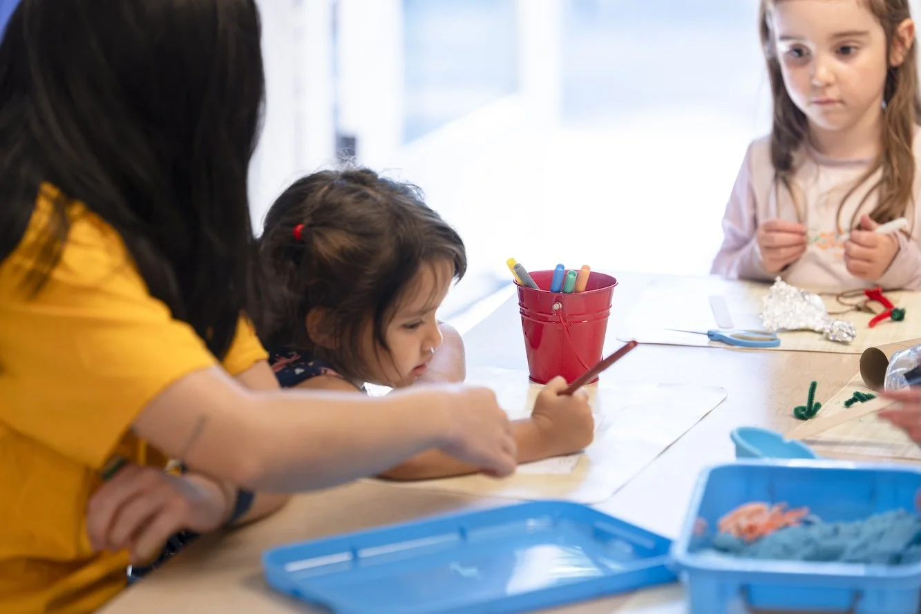 Children and an adult engaging in a craft activity at a table, with craft supplies such as markers, scissors, and pipe cleaners visible.