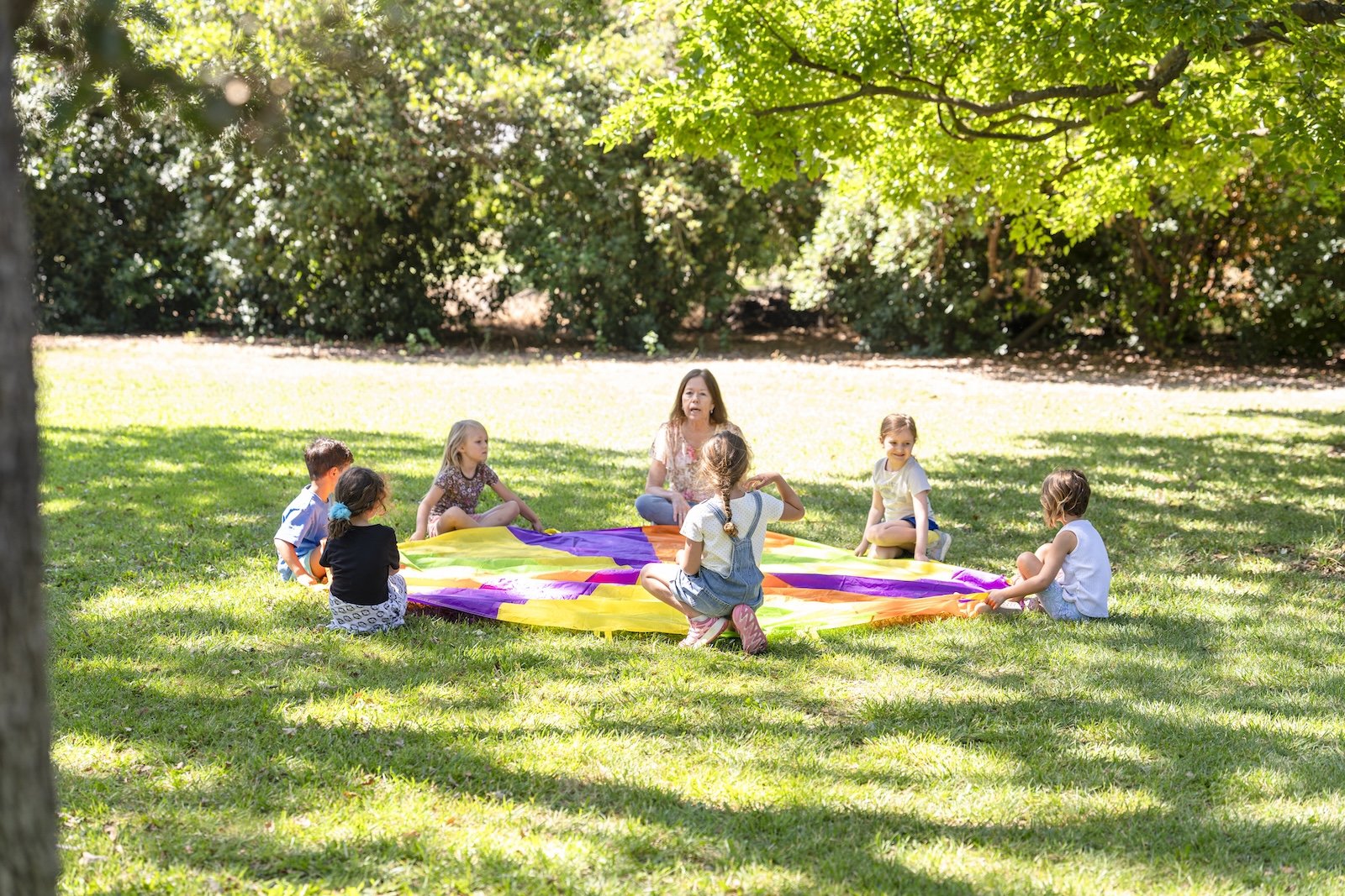 A group of children sitting in a circle outdoors on a grassy field, playing with a rainbow-colored parachute, under a large leafy tree on a sunny day.