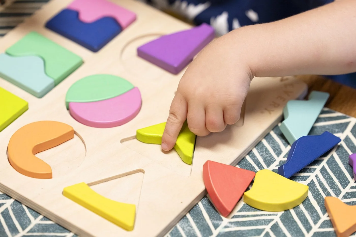 Child's hand placing a yellow wedge into a shape sorter toy with various colorful geometric shapes.