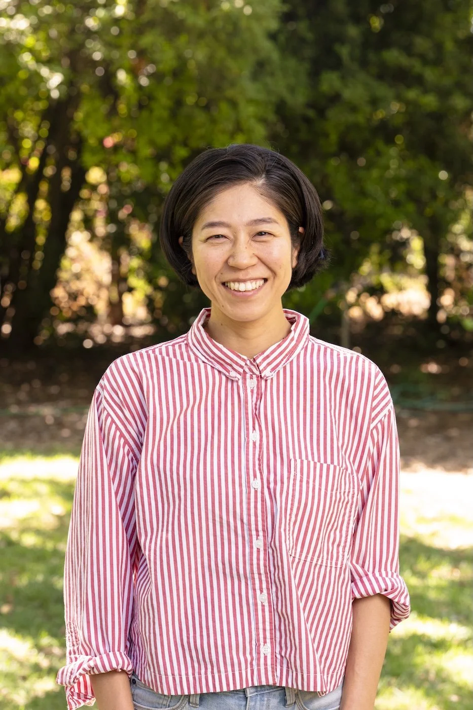 A smiling woman outdoors with a background of trees and sunlight, wearing a red and white striped button-up shirt with rolled-up sleeves.