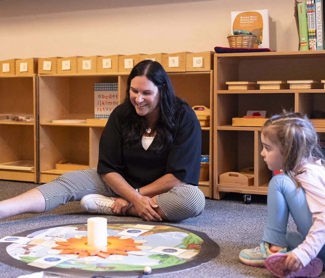 A teacher and a young girl sitting on the carpet in a classroom, engaging in a learning activity centered around a colorful, circular game or puzzle on the floor, with wooden shelves and educational materials in the background.