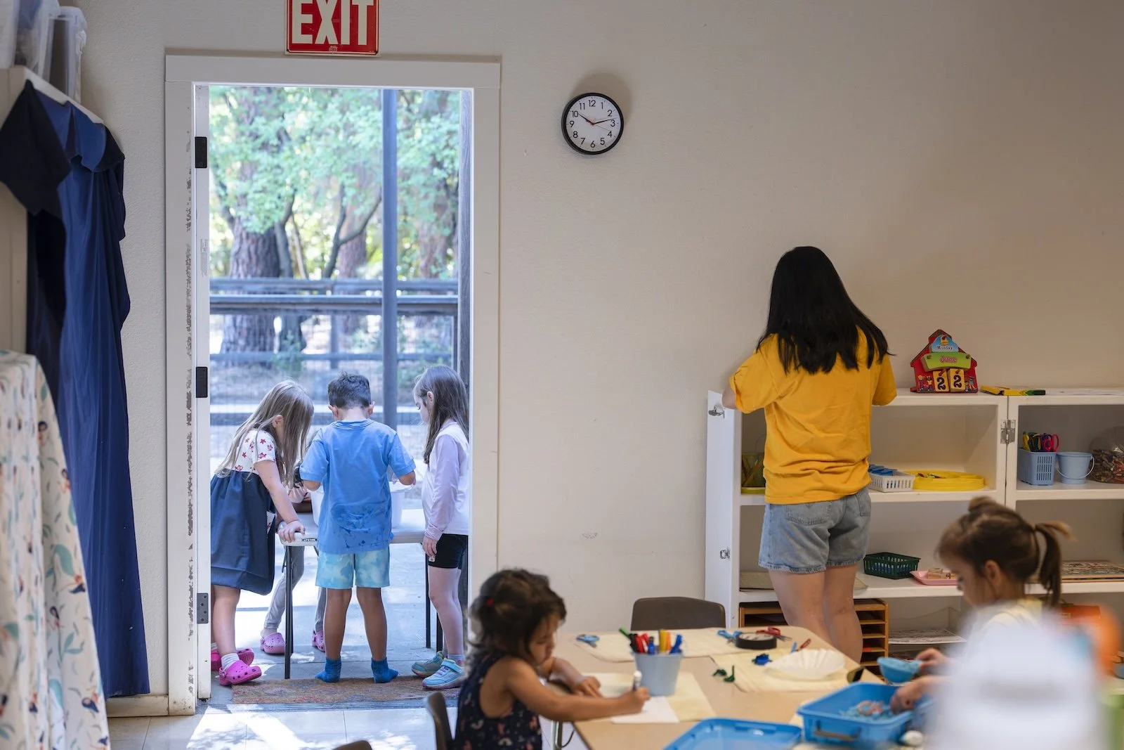 Children inside a classroom with a woman in a yellow shirt near a shelf, and children playing outside through an open door with additional children gathered outside.