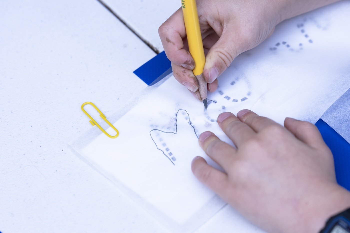 Person drawing a design on tissue paper with a pencil for tattooing, with a paperclip nearby on a white surface.