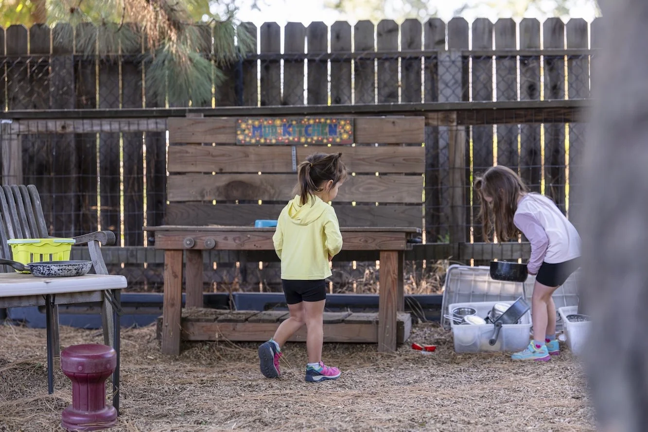 Two young girls playing in a backyard mud kitchen area. One girl is standing, wearing a yellow hoodie and shorts, while the other girl is squatting, wearing a pink top and shorts. There are plastic dishes and utensils in a container and on a table ne