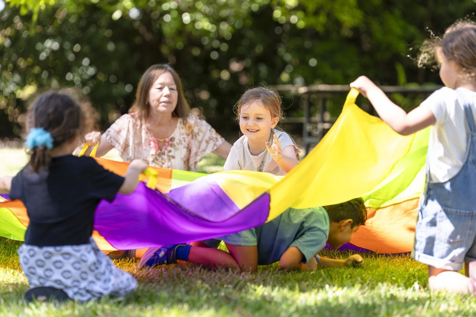Children and an adult playing with a colorful parachute outdoors on grass, surrounded by trees.