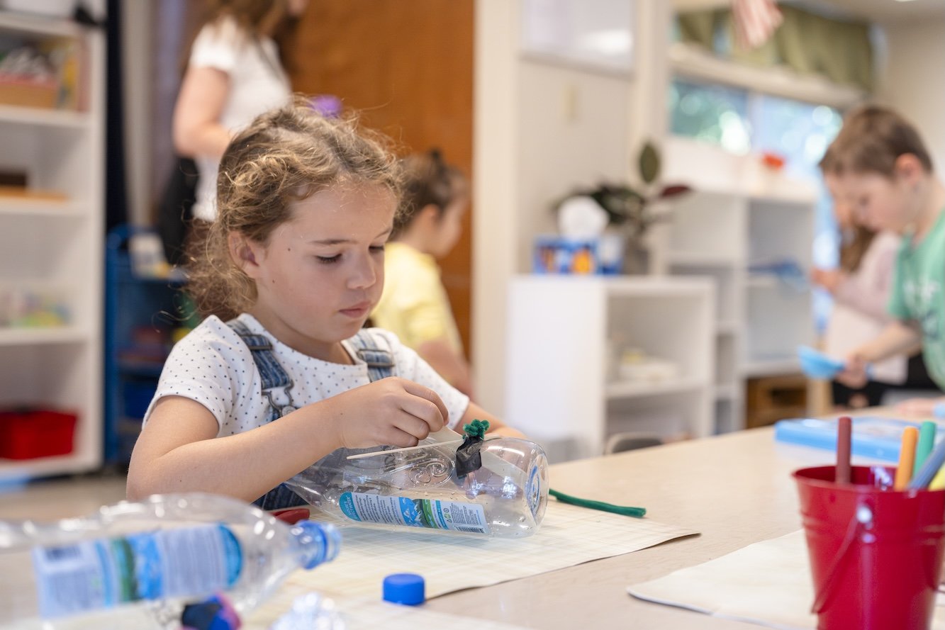 A young girl with curly hair in a white polka-dot shirt and denim overalls working on a craft project at a school table, holding a plastic bottle.